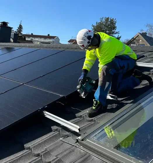 An installer in a helmet and safety harness fixing rails beside finished solar panels on a roof.
