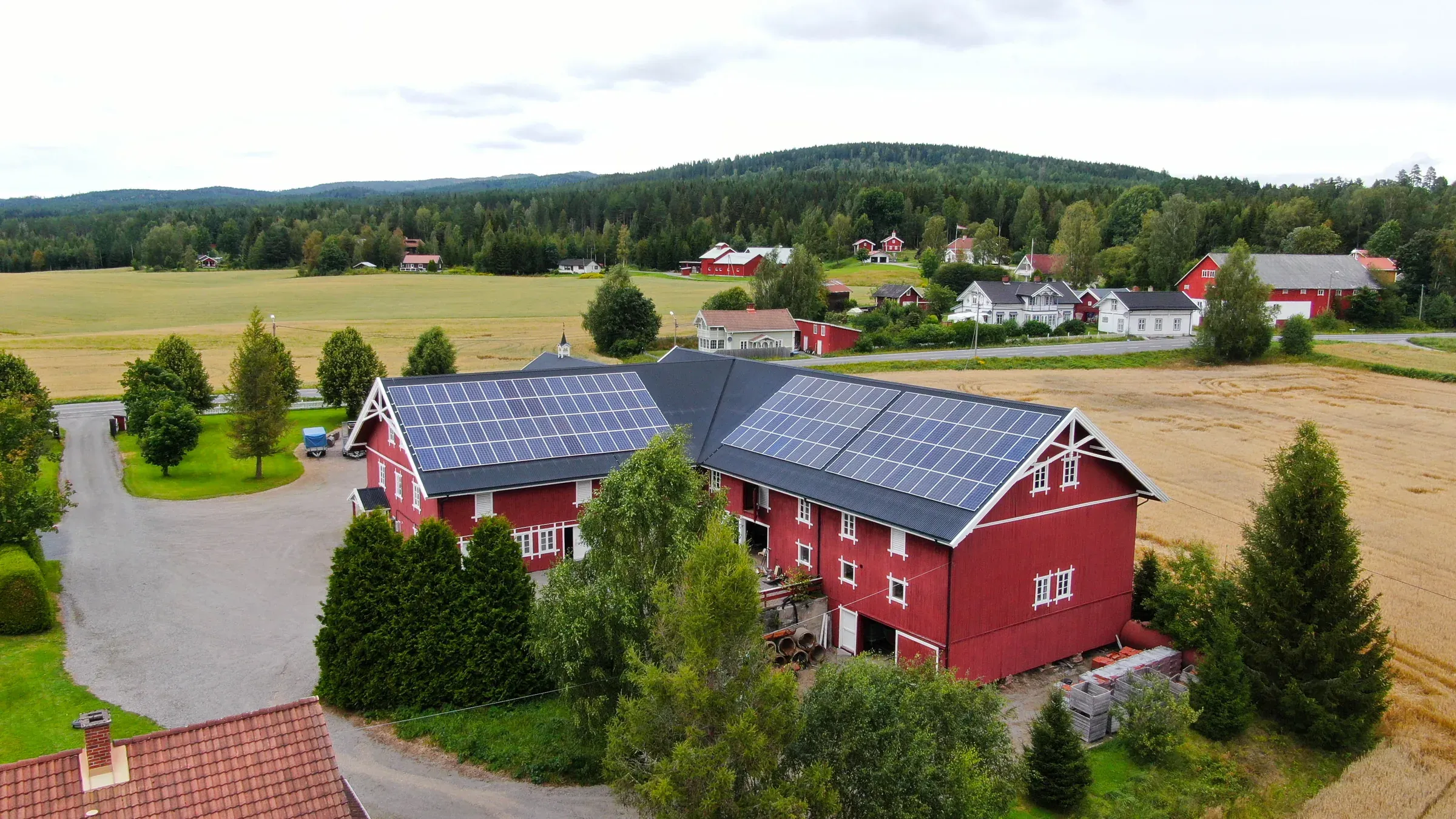 Dronefoto av en stor rød låve med solcellepaneler på to takflater, omgitt av kornåker og skog.