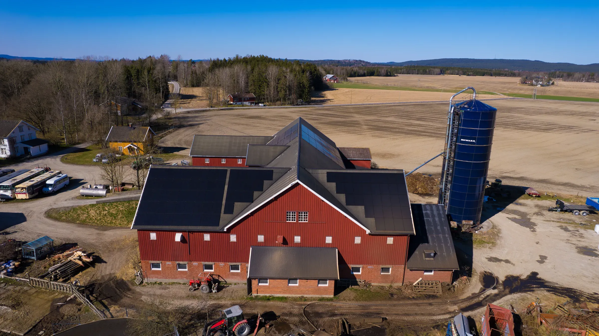 Dronefoto av en rød låve med solcellepaneler på taket, med en silo og jorder rundt.