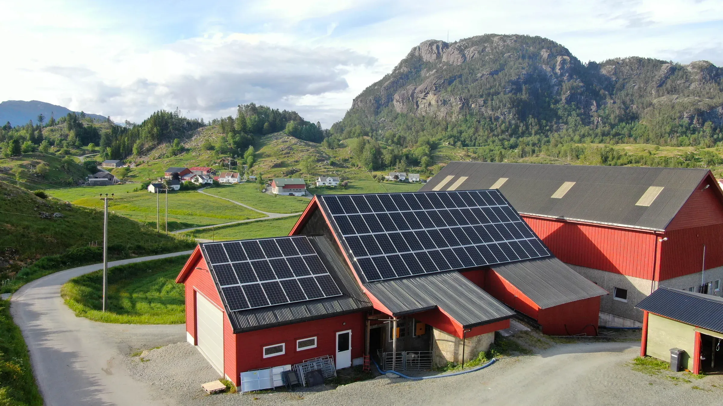 Dronefoto av røde driftsbygninger med solcellepaneler på taket, med grønne jorder og fjell i bakgrunnen.