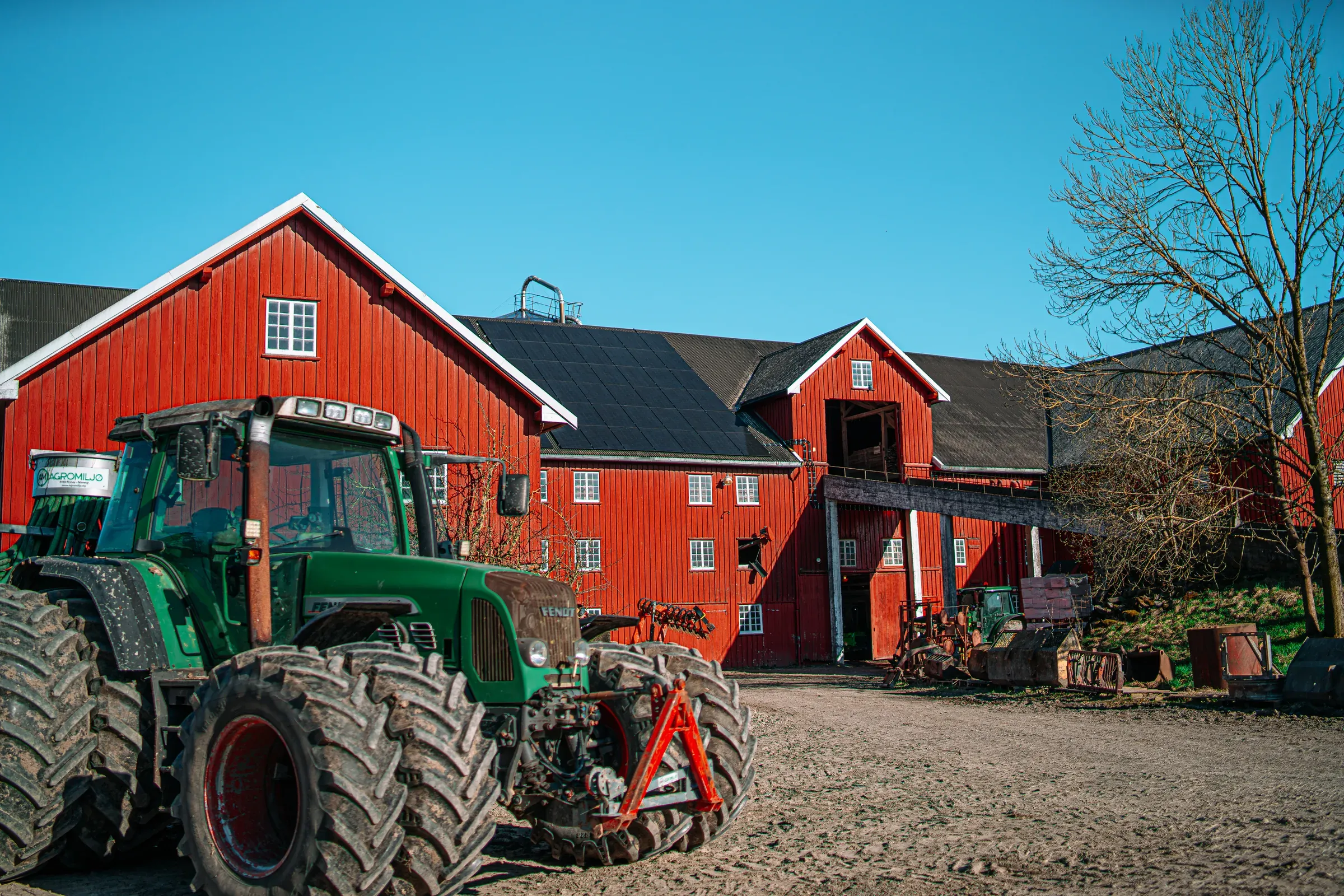 Traktor på gårdsplassen foran en rød låve med solcellepaneler på taket.